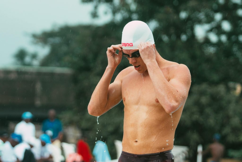 A shirtless man wearing a white swim cap and goggles adjusts his gear poolside, with water dripping from his body. The background features an outdoor setting with blurred figures and trees, evoking a sense of focus and athletic preparation.