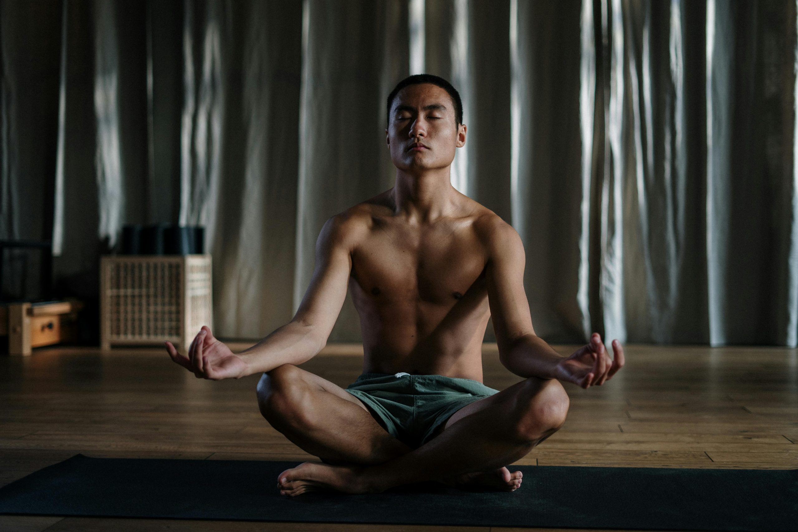 A shirtless man sitting cross-legged on a yoga mat in a meditative pose, in a serene indoor setting with soft lighting and curtains in the background.