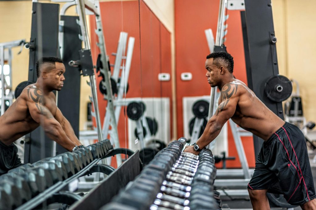 A strong and healthy man with a confident expression, standing in a gym and leaning against a row of dumbbells, gazing into a mirror.