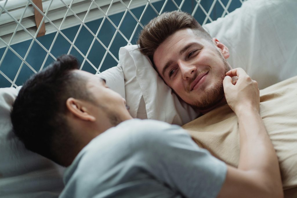 Two men lying in bed together, smiling warmly at each other, sharing a tender and intimate moment.