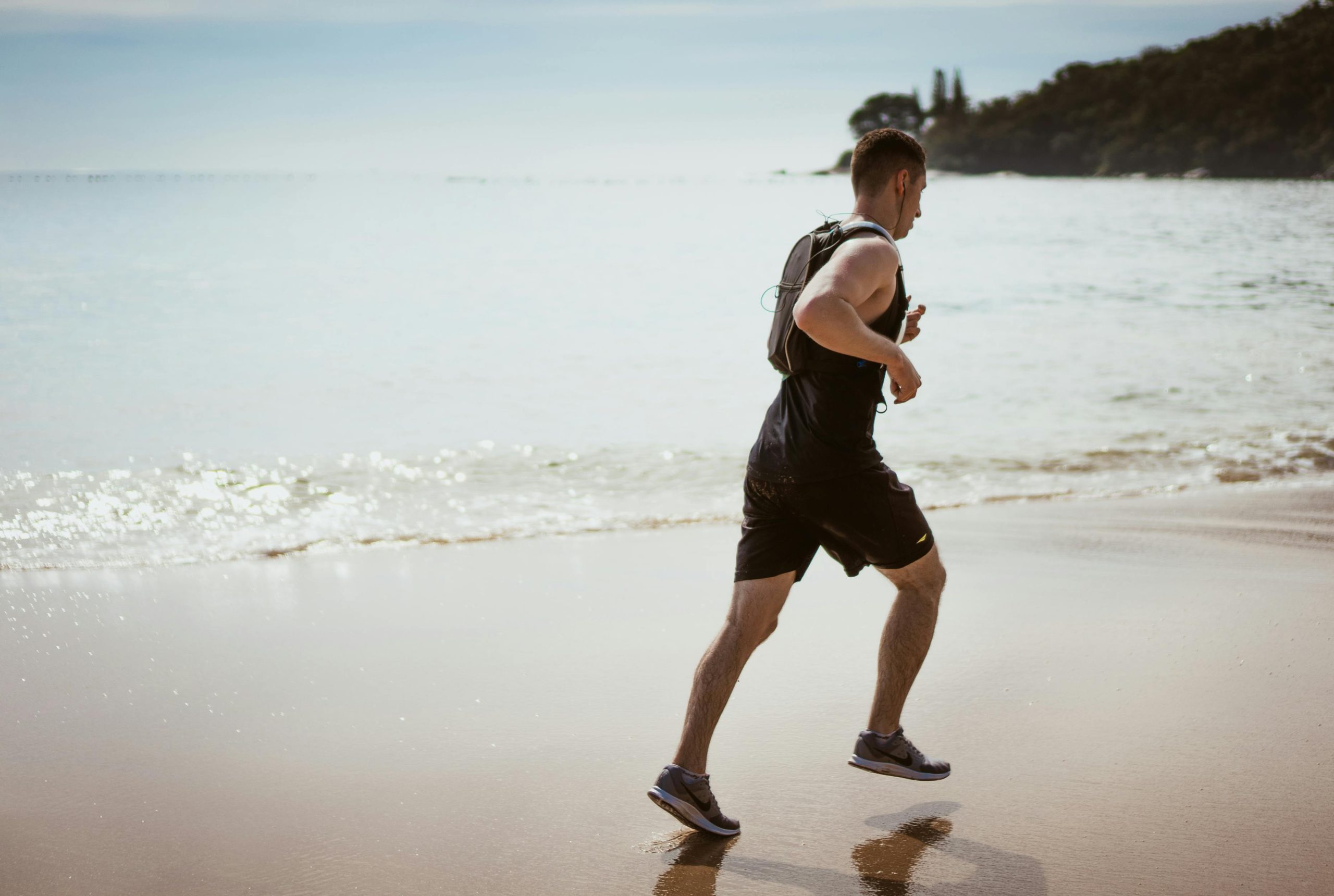 A fit man jogging along a sandy beach under clear skies, wearing athletic gear and a hydration pack, with a calm sea and distant greenery in the background.