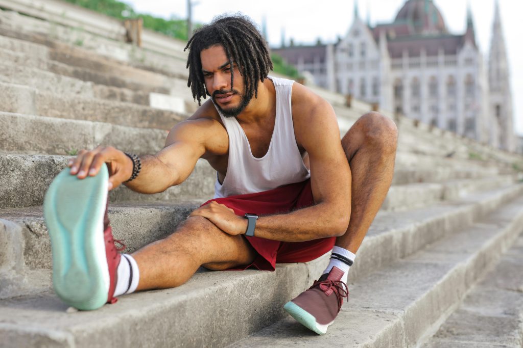 A fit young man with stretches on outdoor steps, wearing a white tank top, red shorts, and athletic shoes.