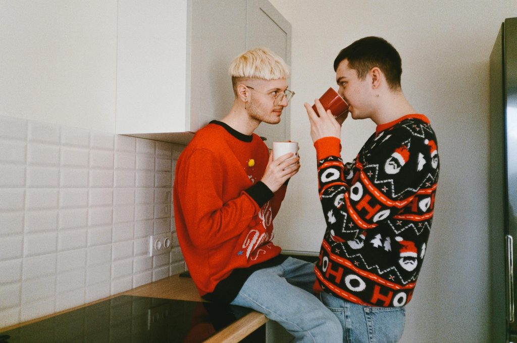 Two young men in festive sweaters enjoying warm drinks together in a cozy kitchen setting, sharing a moment of connection and warmth.