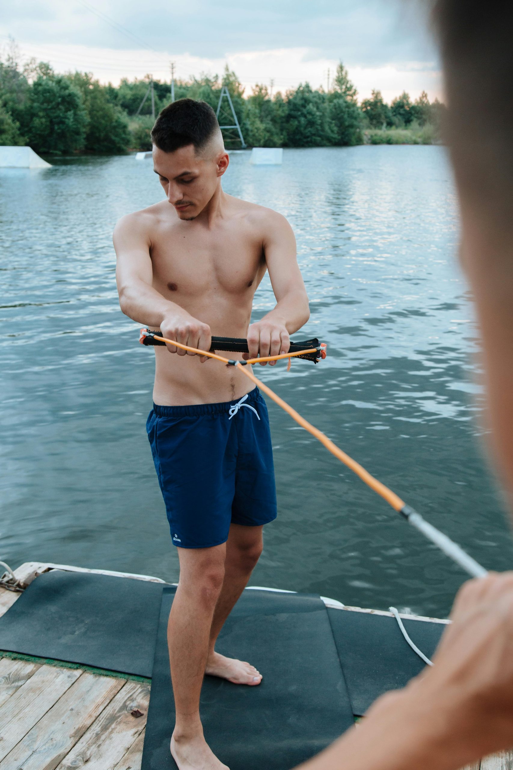 A young, fit man standing on a dock by a serene lake, shirtless and wearing blue swim shorts, as he grips a waterski handle, preparing for an activity on the water.