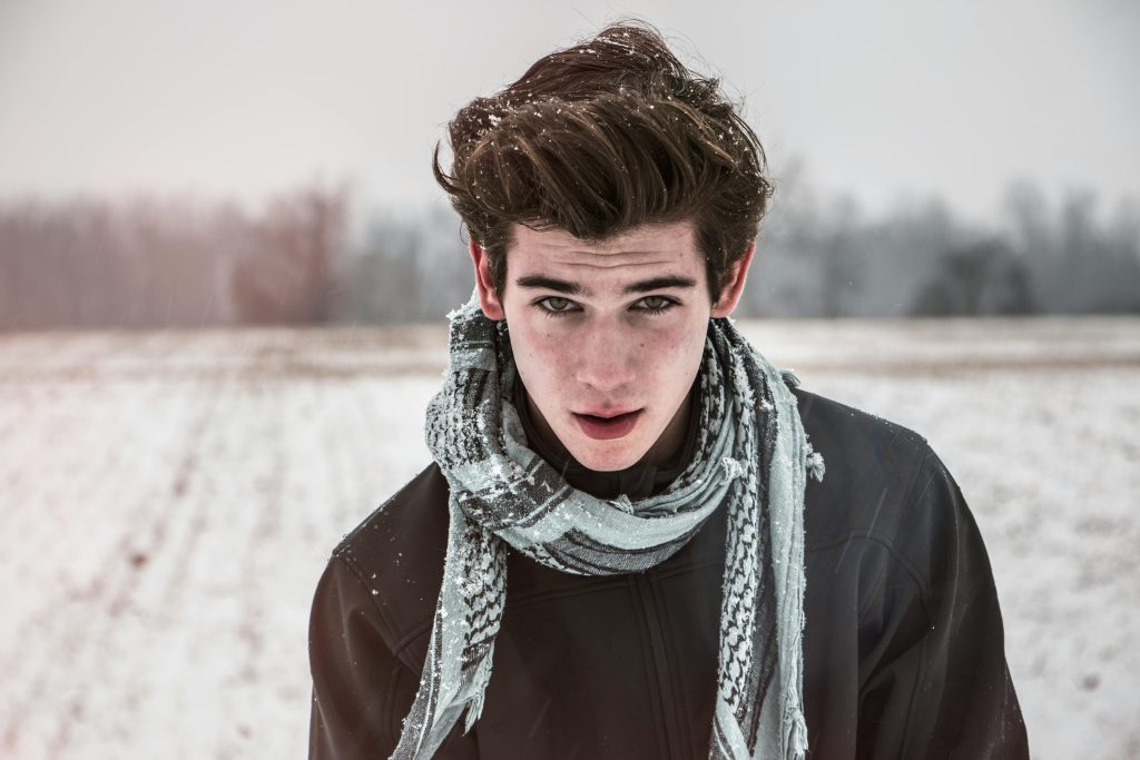 A young man with dark hair and a stylish scarf stands in a snowy field, gazing into the camera with a confident expression. Snowflakes rest on his hair and clothing, adding to the wintery atmosphere.