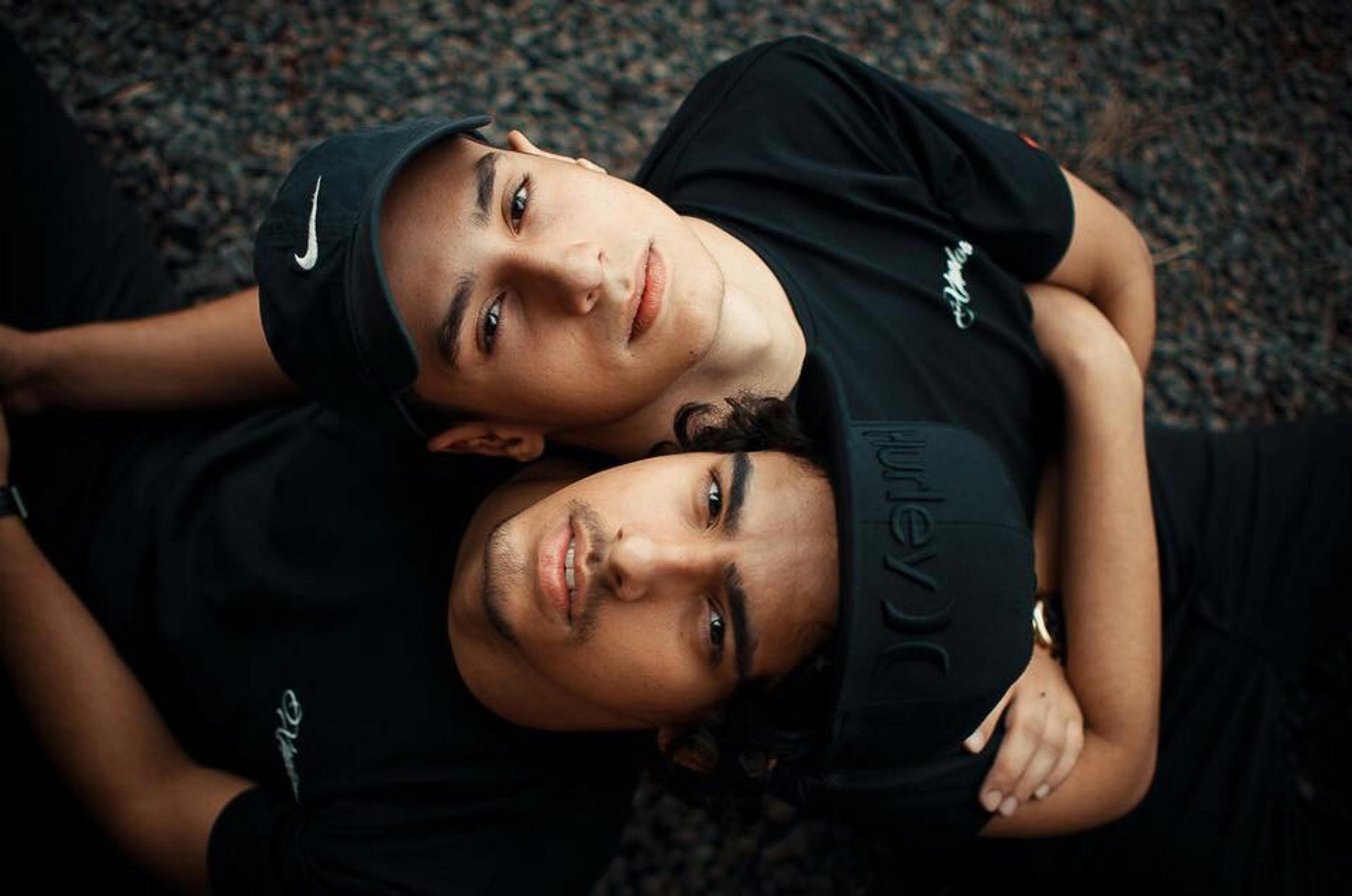 Two young men in black shirts and caps lying on the ground, leaning against each other, sharing a supportive and intimate moment.